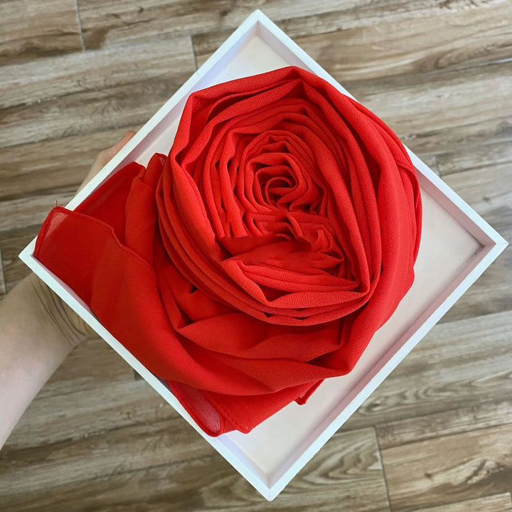 Hand holding a white square tray with a neatly folded red chiffon fabric resembling a rose, wooden floor background