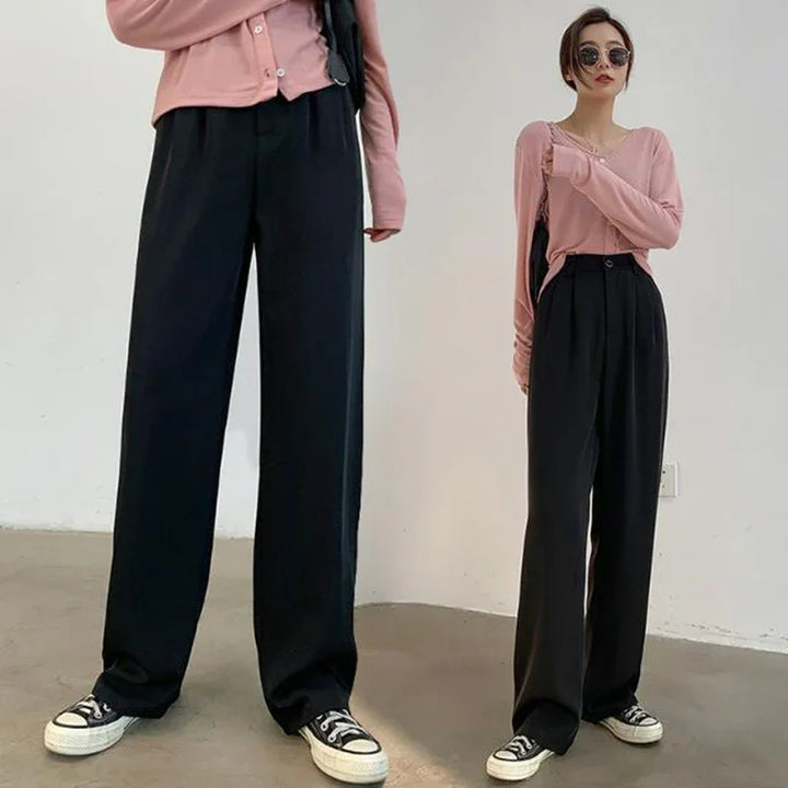 Woman in pink long-sleeve top, black wide-leg pants, and black sneakers against a plain indoor wall