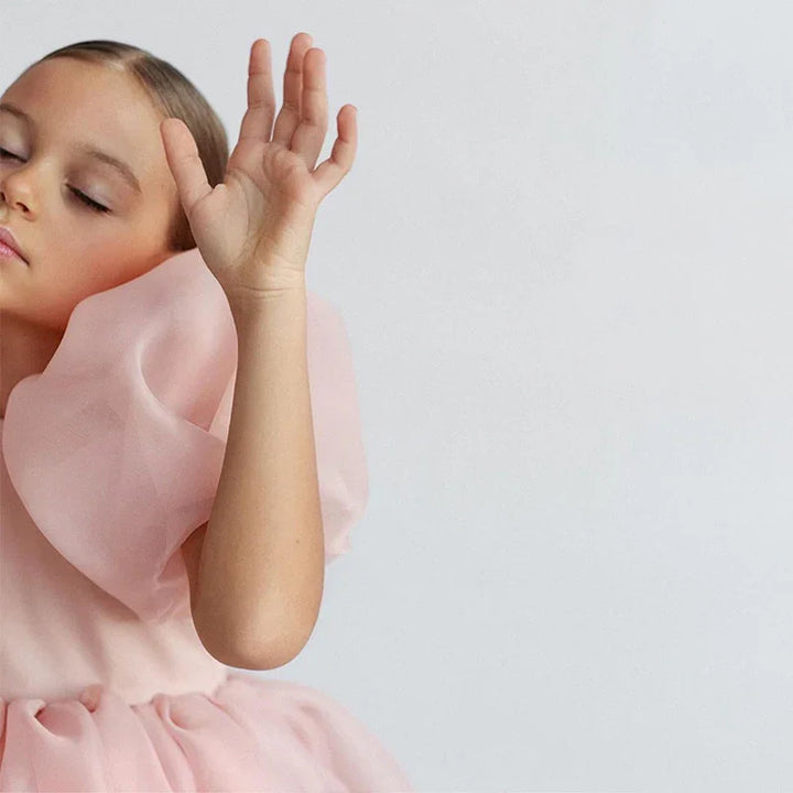 Young girl in a soft pink dress with large ruffles, posing gracefully with eyes closed against a plain background