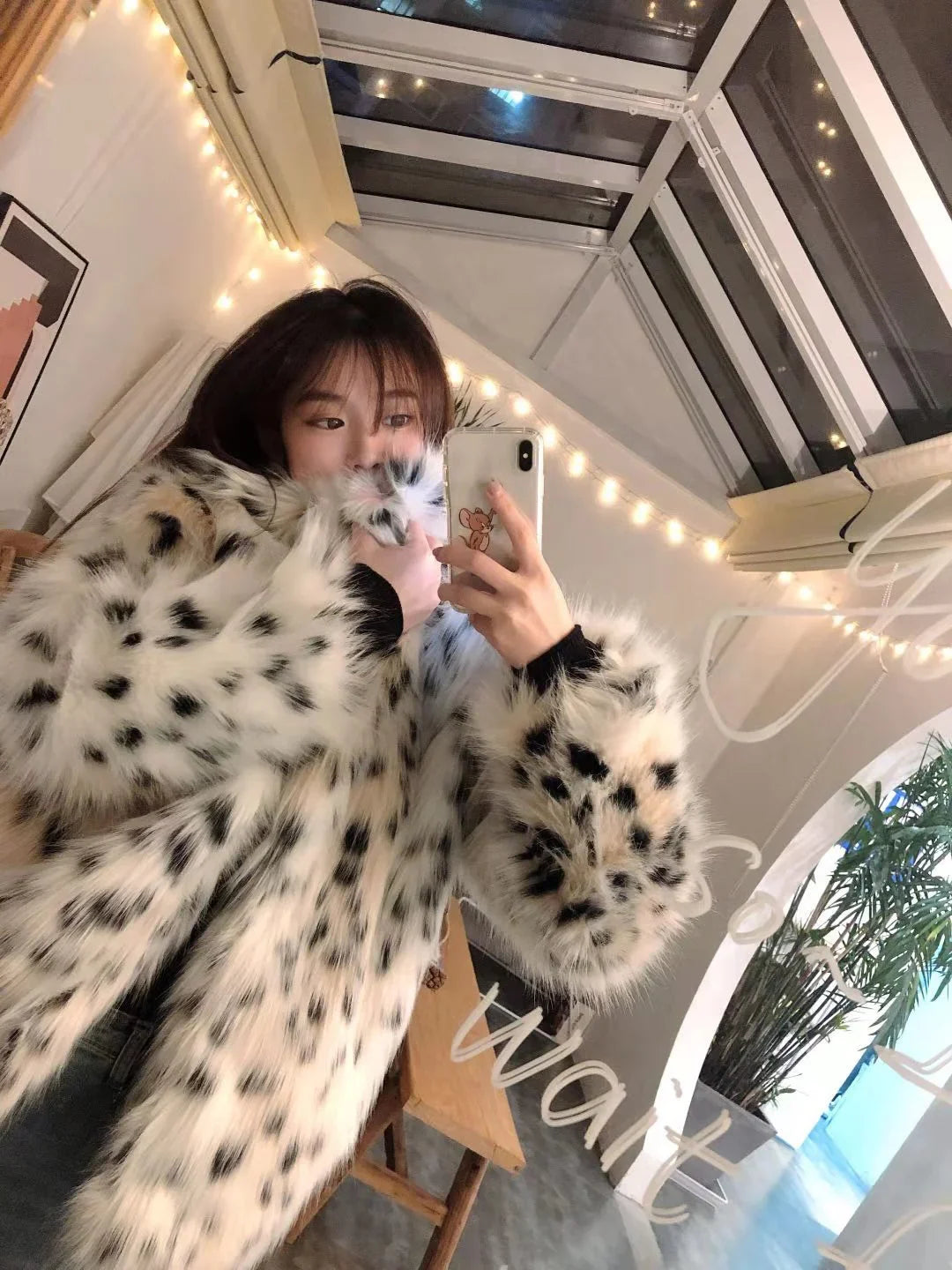 Young woman taking mirror selfie wearing spotted faux fur coat in cozy room with string lights