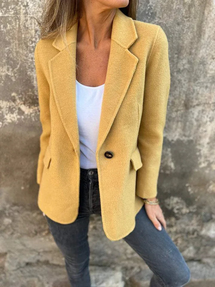 Woman wearing mustard yellow wool blazer, white top, and dark jeans against textured wall