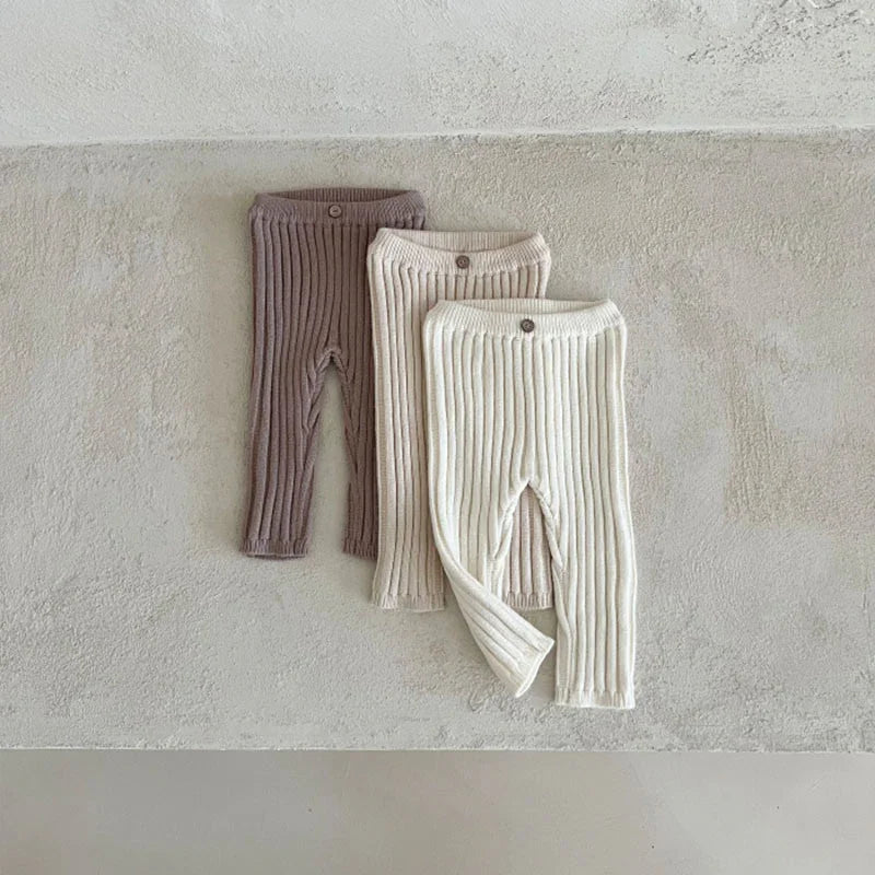 Three ribbed knit baby leggings in brown, beige, and cream colors laid flat on textured surface
