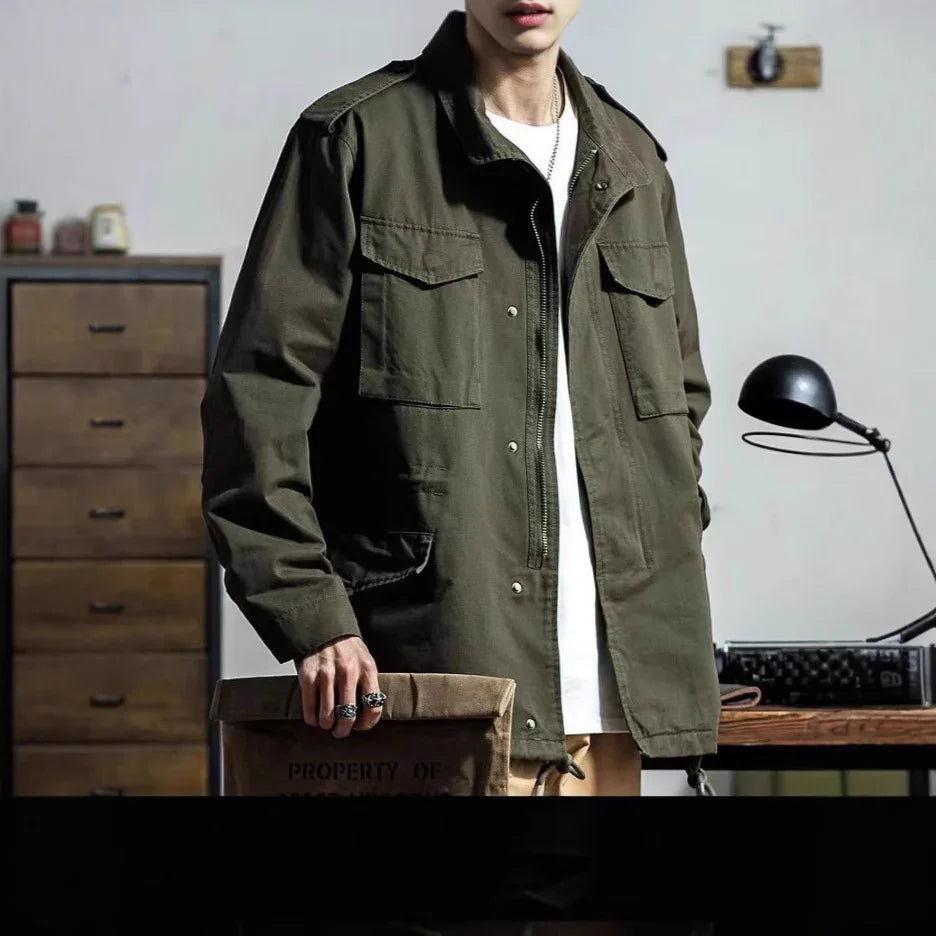 man wearing olive green military-style jacket over white shirt holding brown canvas bag indoors