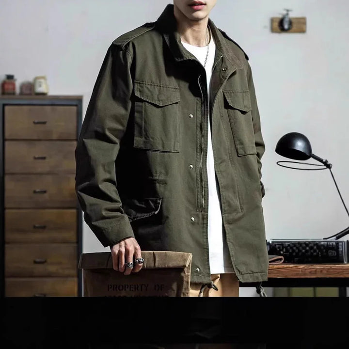 man wearing olive green military-style jacket over white shirt holding brown canvas bag indoors