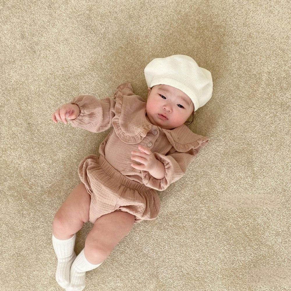 Asian baby lying on beige carpet wearing beige romper, white socks, and cream beret hat