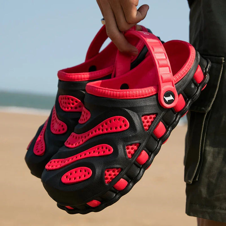 Hand holding black and red ventilated casual slip-on sandals with adjustable heel straps at the beach