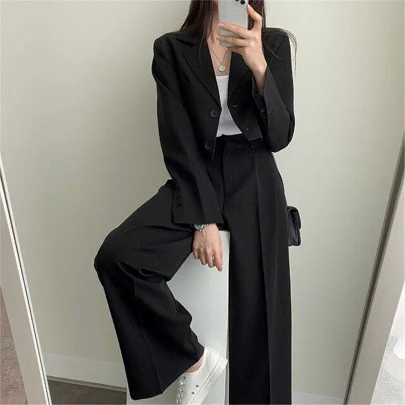 Woman wearing black blazer and wide-leg trousers with white top and sneakers, taking mirror selfie