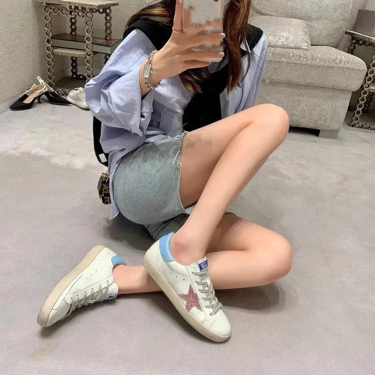 Person sitting on carpet floor wearing white sneakers with pink star and light blue accents, denim shorts, and light blue shirt in stylish indoor setting
