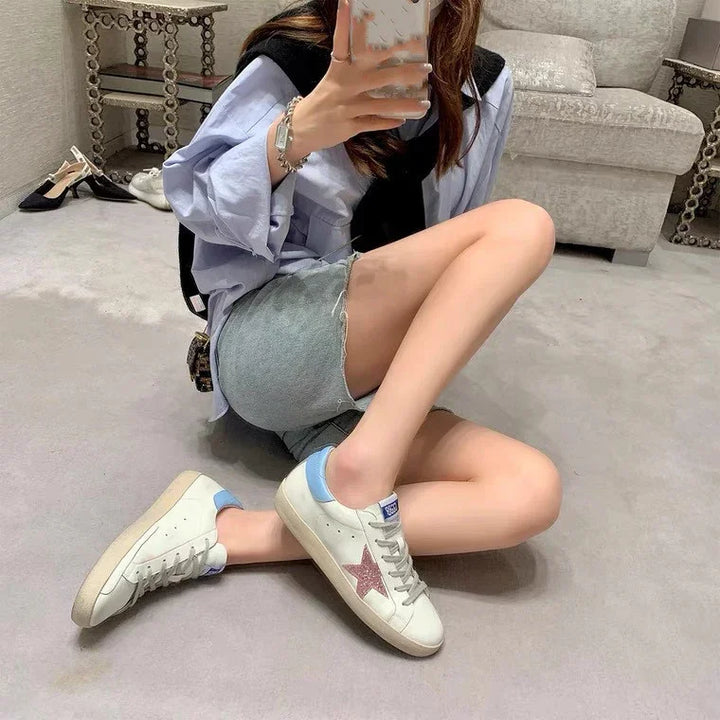 Person sitting on carpet floor wearing white sneakers with pink star and light blue accents, denim shorts, and light blue shirt in stylish indoor setting
