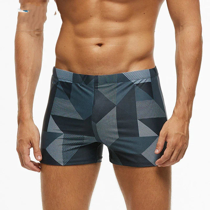 Male model wearing geometric patterned black and gray swim trunks showcasing fitness and swimwear fashion