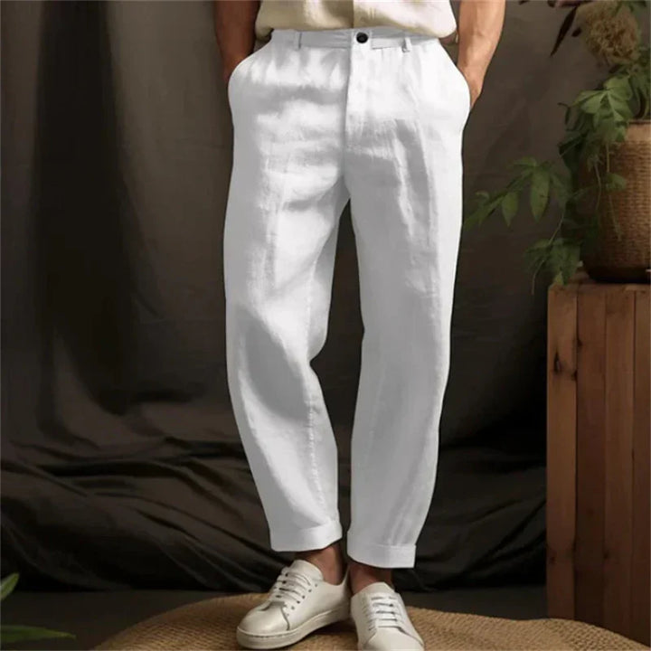 Man wearing white casual linen pants and white sneakers standing indoors with plants and wood decor