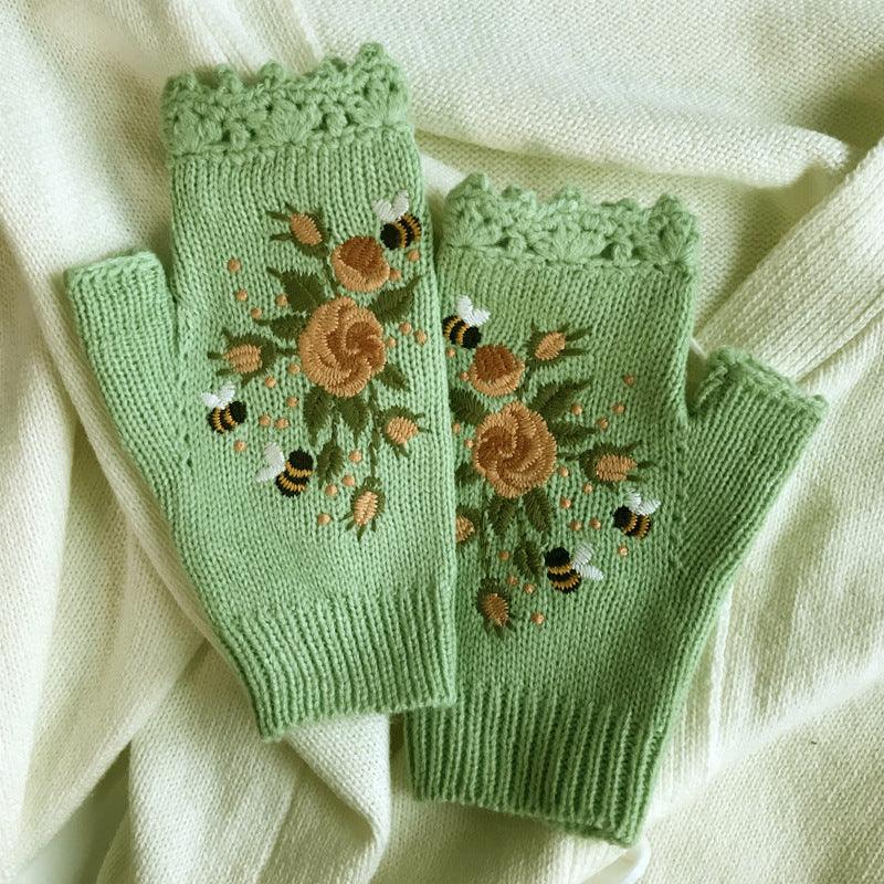 Green knitted fingerless gloves with floral and bee embroidery, laid on cream fabric