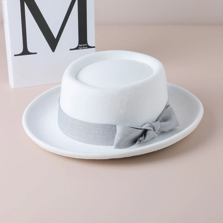 White fedora hat with a light gray ribbon and bow, placed on a beige surface with a decorative book in the background