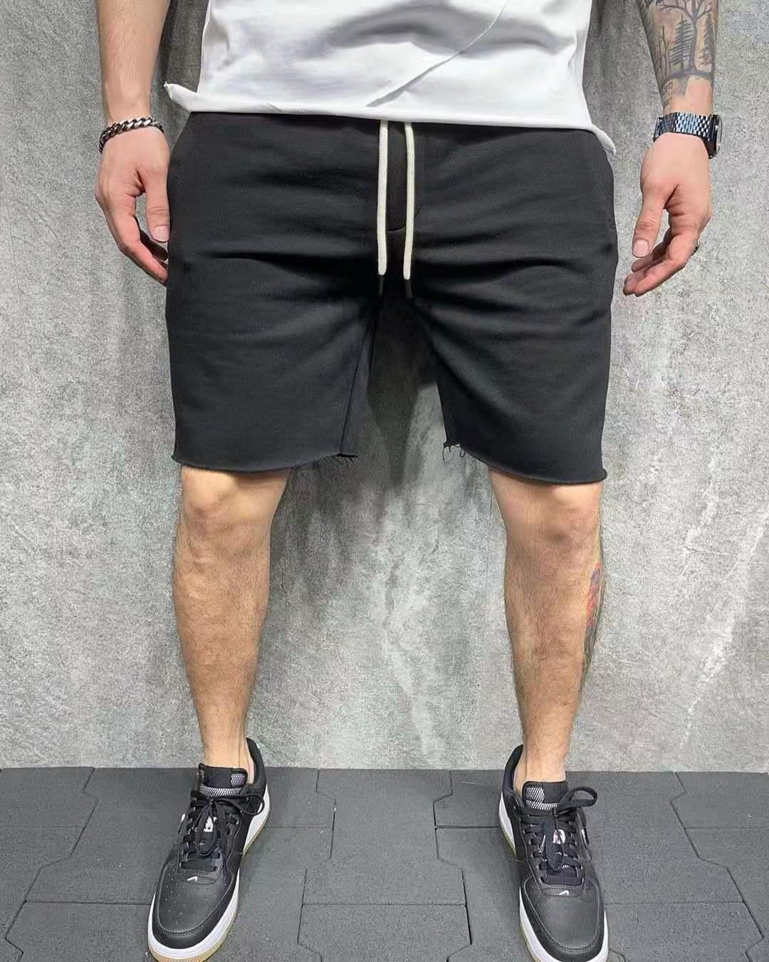 Man wearing black casual shorts with white drawstring and black sneakers against gray wall