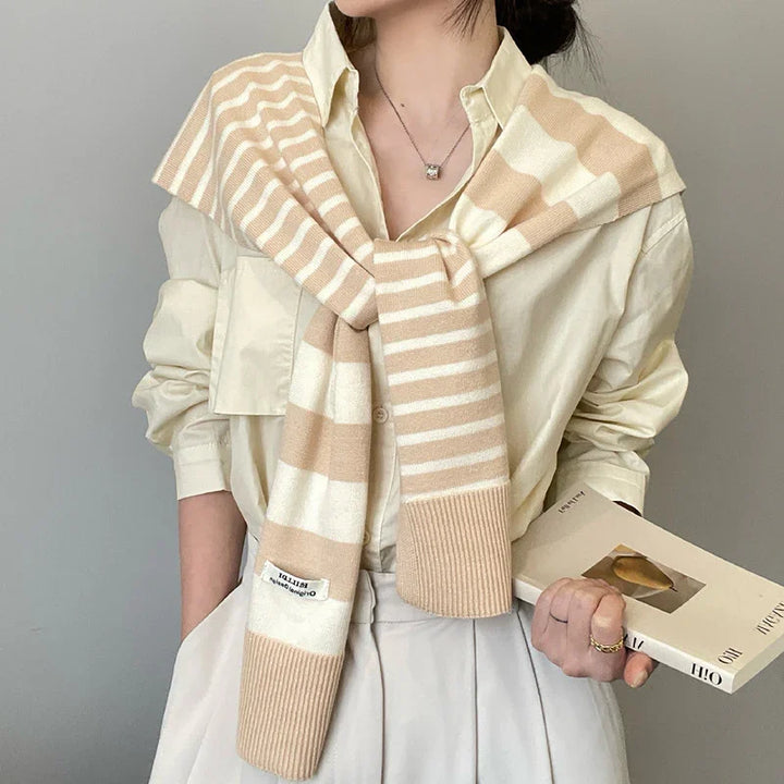 Woman wearing beige striped knit scarf over cream blouse holding art book against gray wall