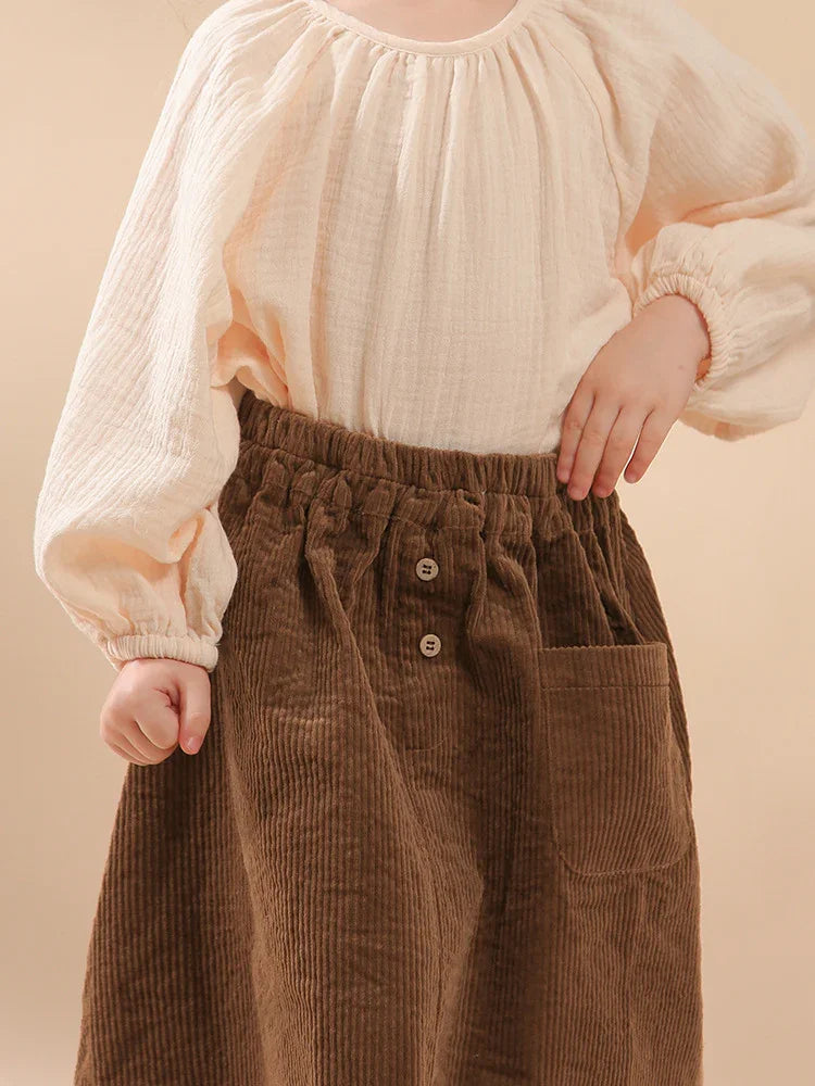 Child wearing cream-colored textured blouse with long sleeves and brown corduroy skirt with buttons and pocket