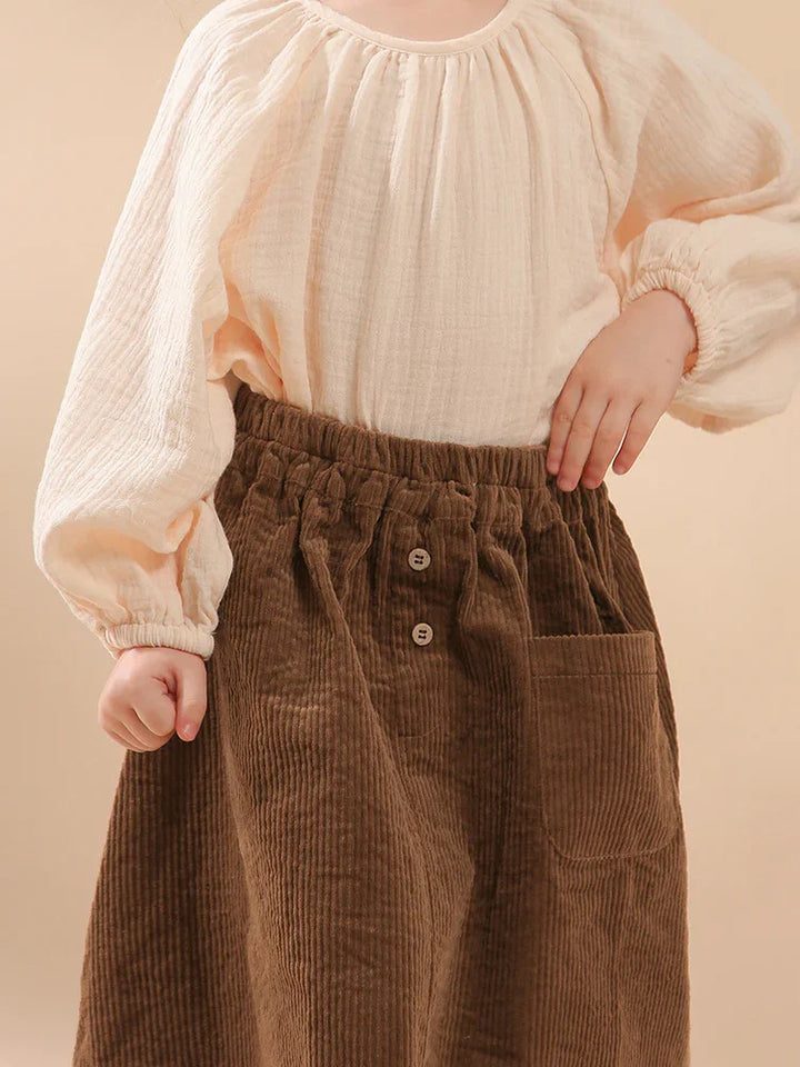 Child wearing cream-colored textured blouse with long sleeves and brown corduroy skirt with buttons and pocket