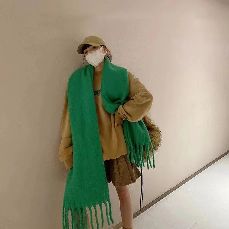Woman in beige oversized sweater and cap with large green scarf and brown pleated skirt in minimalist hallway