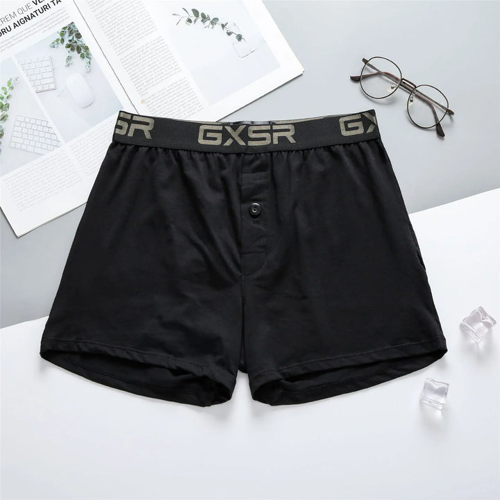 Black men's boxer shorts with GXSR logo waistband, button fly, placed on white and gray background with glasses and magazine