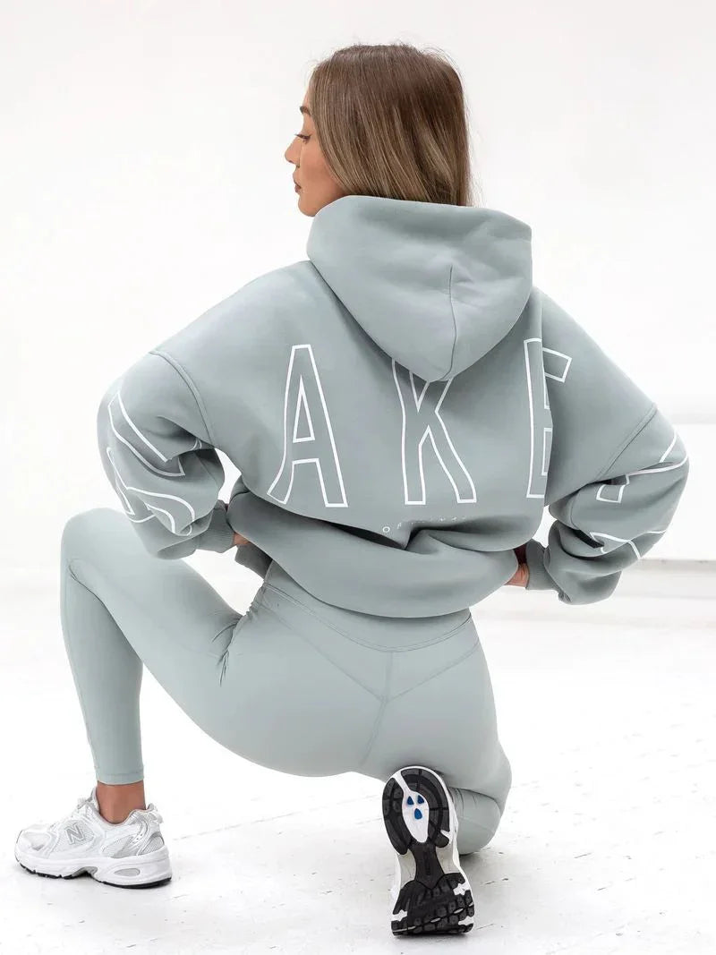 Woman in gray hoodie with large white letters and matching leggings kneeling in white studio