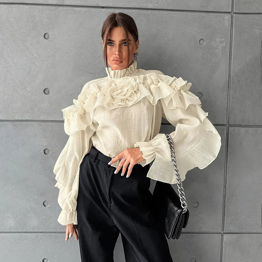 Woman wearing cream ruffled blouse with puff sleeves and black pants posing against gray concrete wall