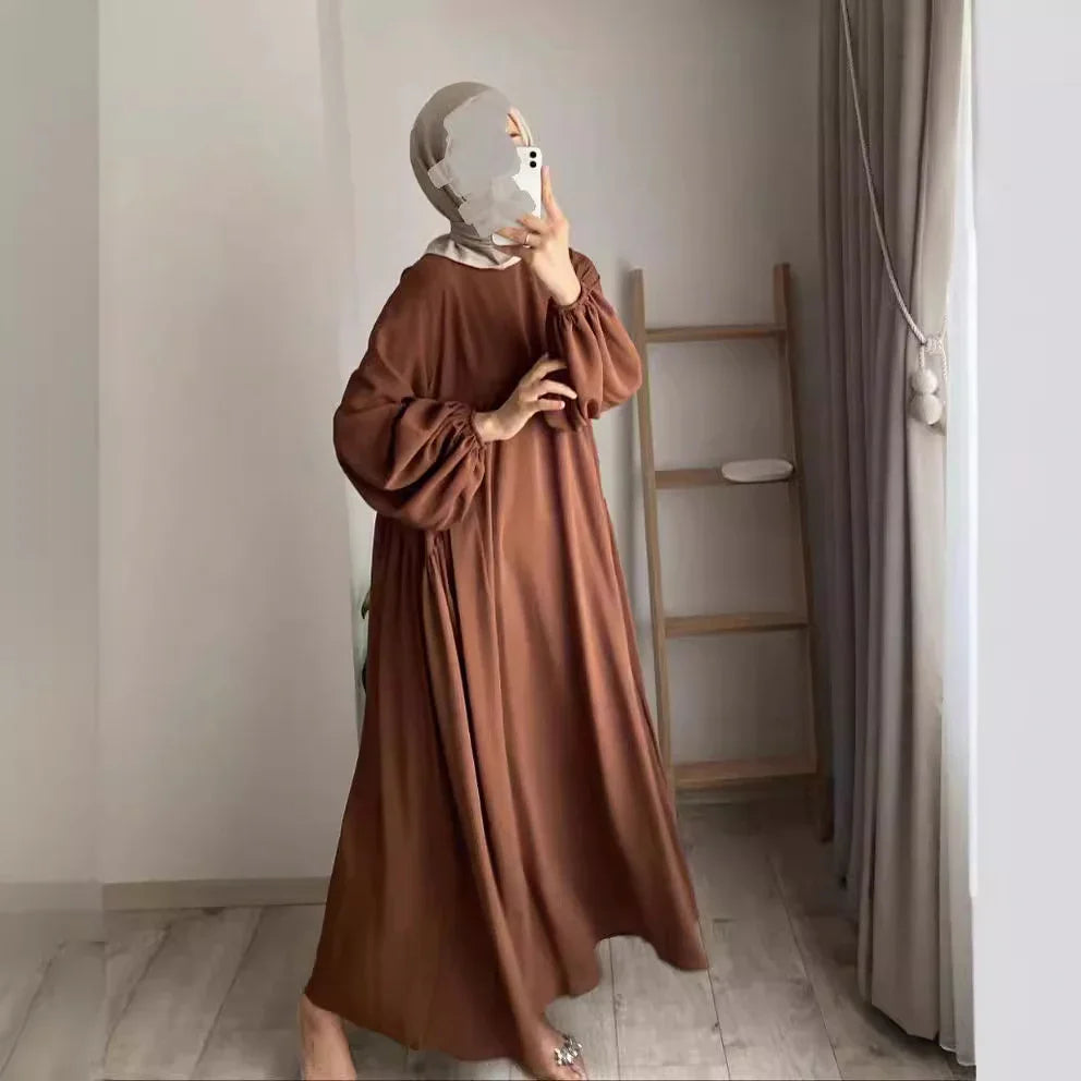 Woman in brown loose dress with puff sleeves and beige hijab taking mirror selfie indoors