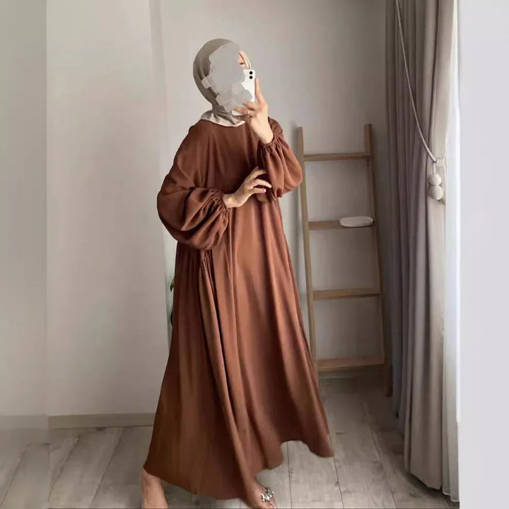 Woman in brown loose dress with puff sleeves and beige hijab taking mirror selfie indoors