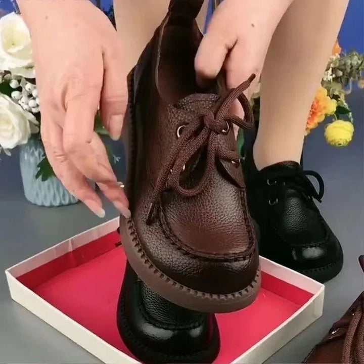 Close-up of hands holding brown leather lace-up shoe over black shoe in a red shoe box with flowers in the background