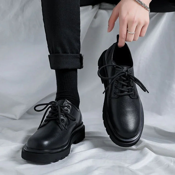 Stylish black leather lace-up shoes worn with black socks and pants, held by a hand with silver rings and bracelet