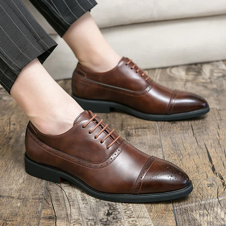Men's brown leather dress shoes with brogue detailing worn with pinstriped trousers on wooden floor