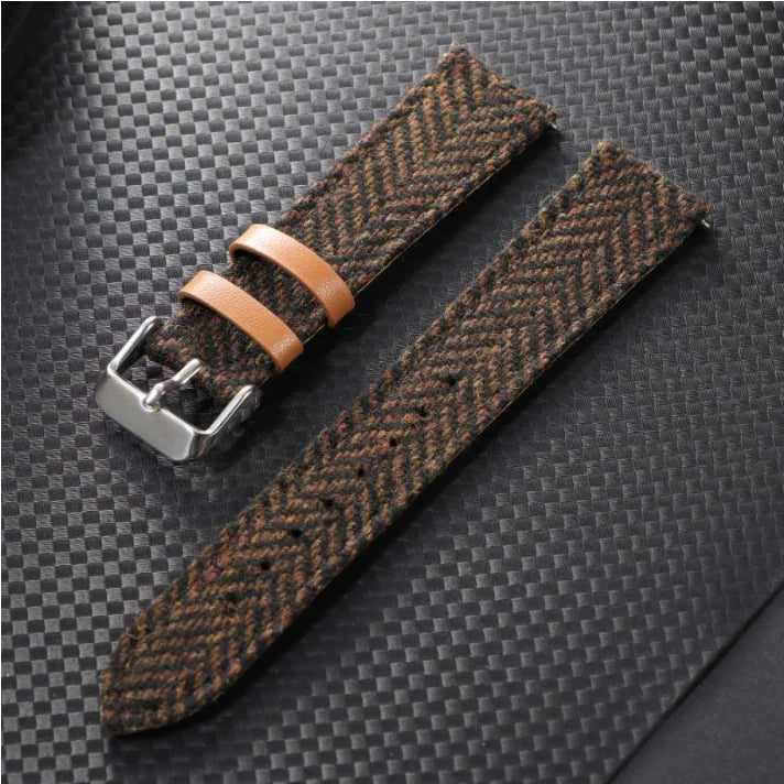 Brown herringbone fabric watch strap with tan leather loops and silver buckle on black textured surface