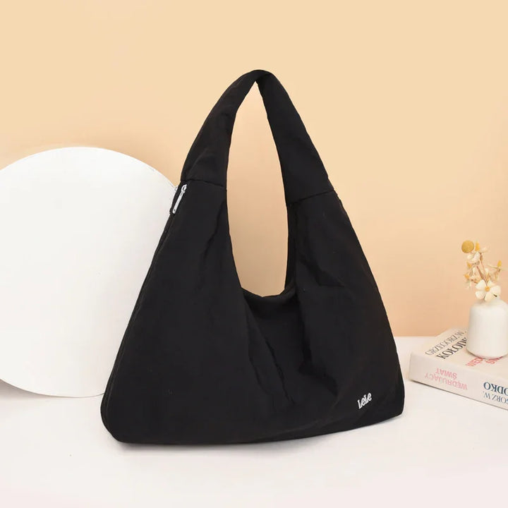 Black triangular shoulder bag with zipper and Lele logo on beige background with books and vase
