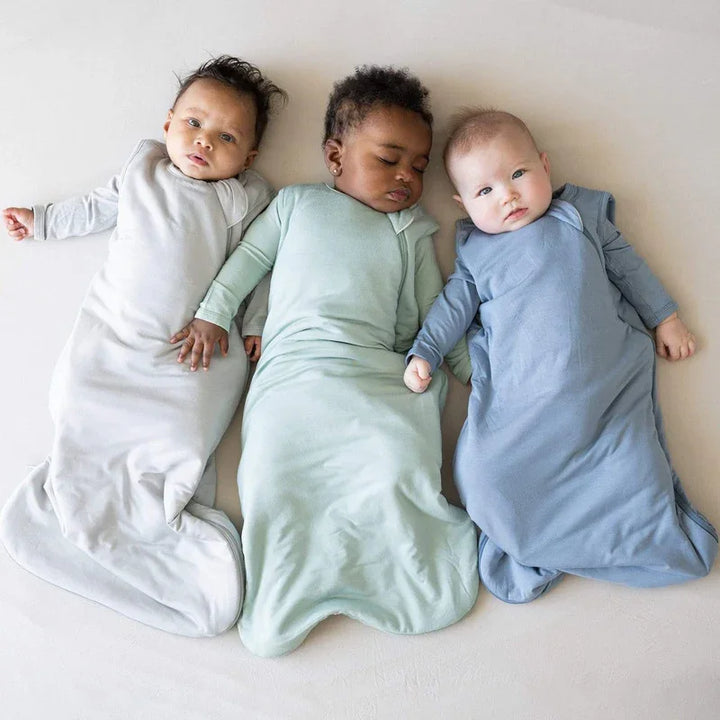 Three babies lying on a beige surface wearing light gray, mint green, and blue wearable blankets