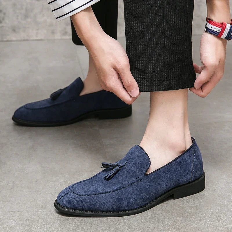 Person wearing navy blue suede tassel loafers and pinstriped black pants adjusting pant hem on gray floor