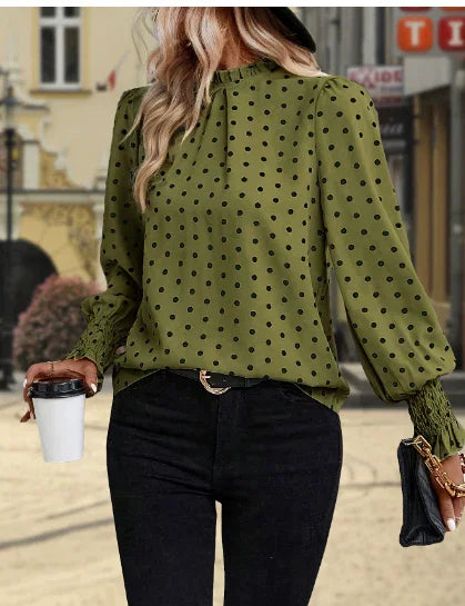 Woman wearing olive green polka dot blouse with puff sleeves and black pants holding coffee cup and clutch