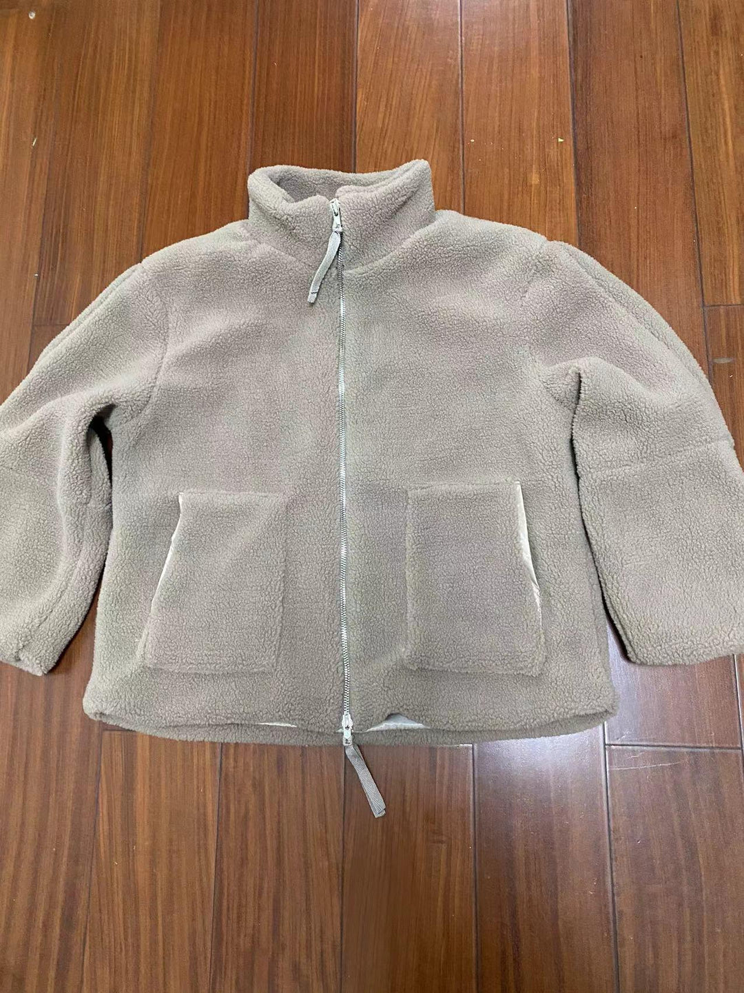 Beige fleece zip-up jacket with two front pockets, laid flat on wooden floor