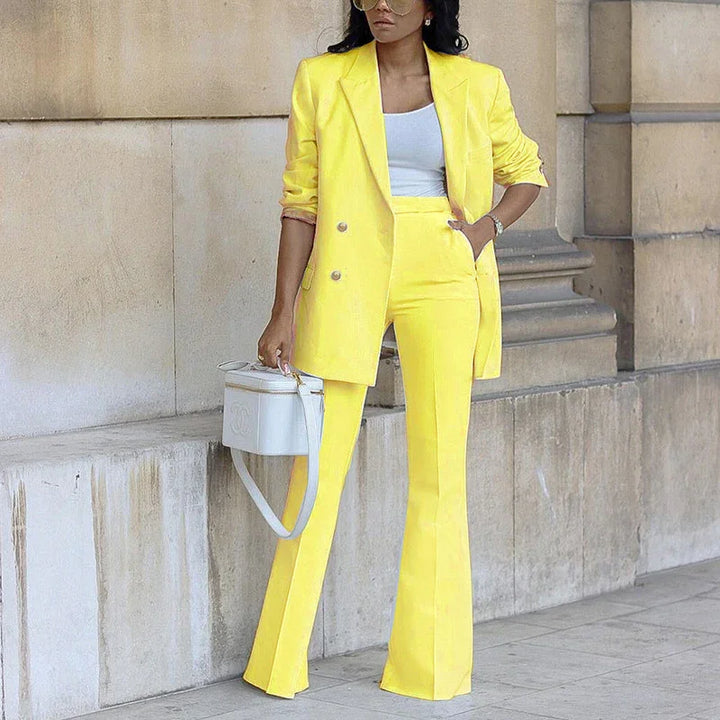 Woman in bright yellow pantsuit with white top and white handbag, posing against stone wall