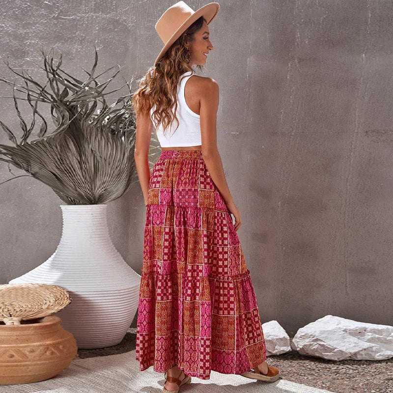 Woman wearing beige hat, white sleeveless top, and patterned red maxi skirt standing indoors by large white vase and decorative elements