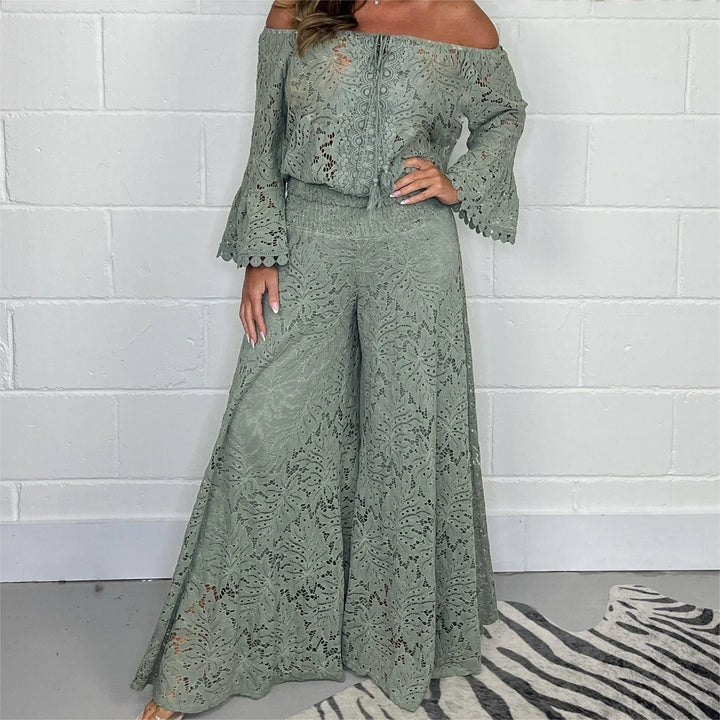 Woman wearing sage green lace off-shoulder top and wide-leg pants set against white brick wall