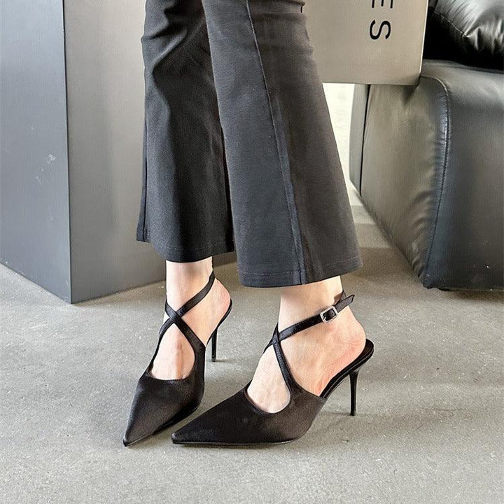 Woman wearing black pointed-toe stiletto heels with ankle straps and cropped black pants indoors