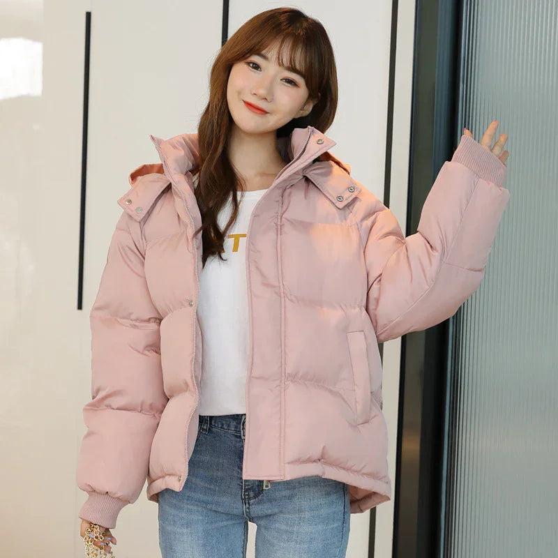 Smiling woman wearing light pink puffer jacket with hood, white tee, and blue jeans indoors