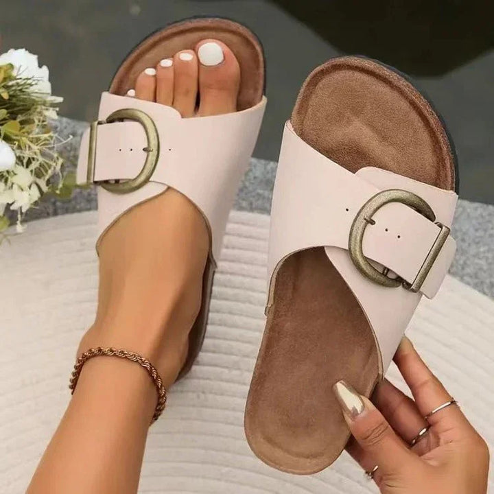 White buckle slide sandals on foot with white pedicure and gold anklet, hand holding one sandal