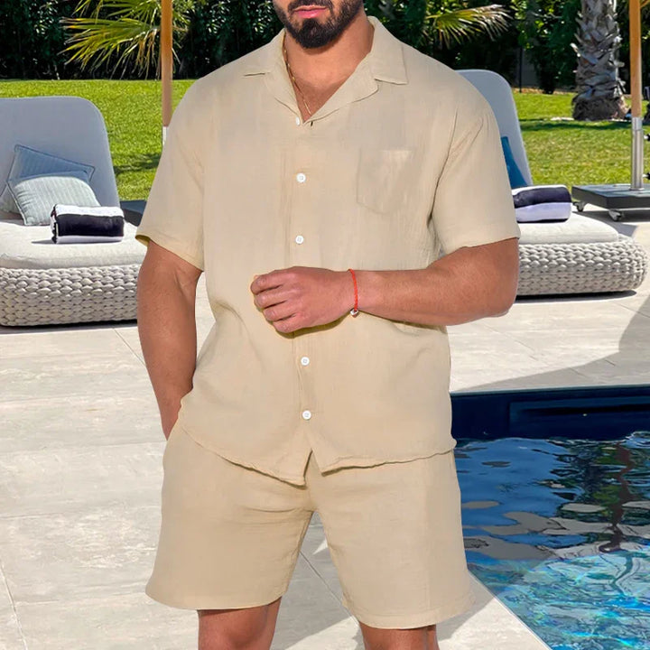 Man wearing beige short-sleeve button-up shirt and matching shorts standing by poolside lounge chairs