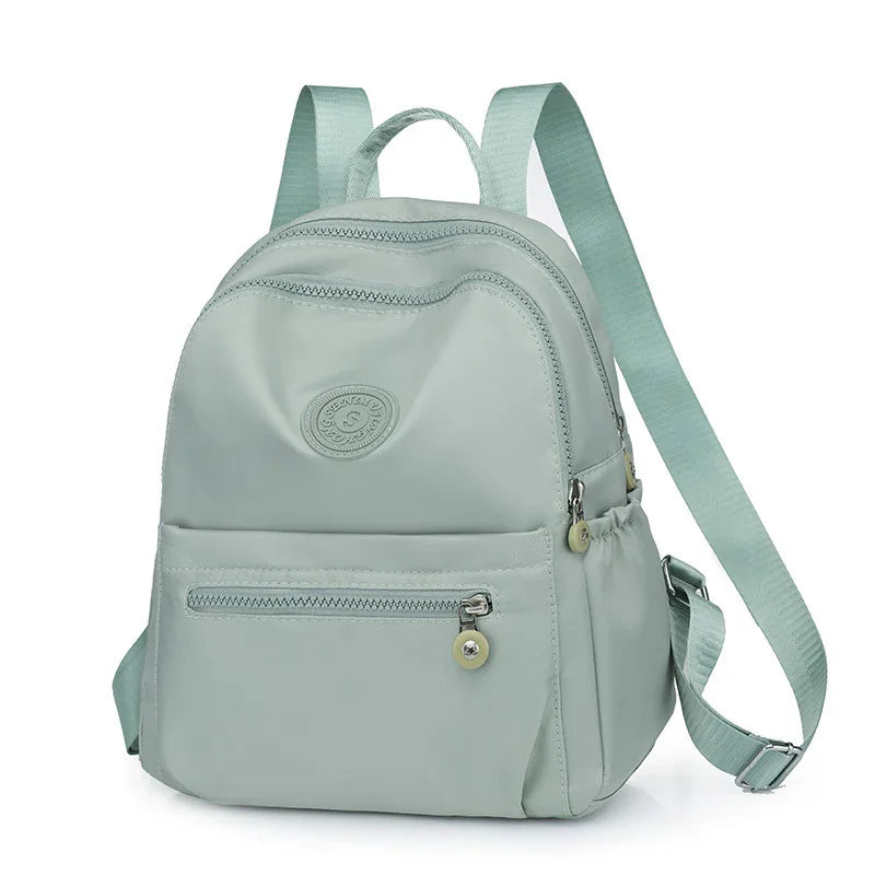 Light gray backpack with front zipper pocket, adjustable straps, and circular logo badge