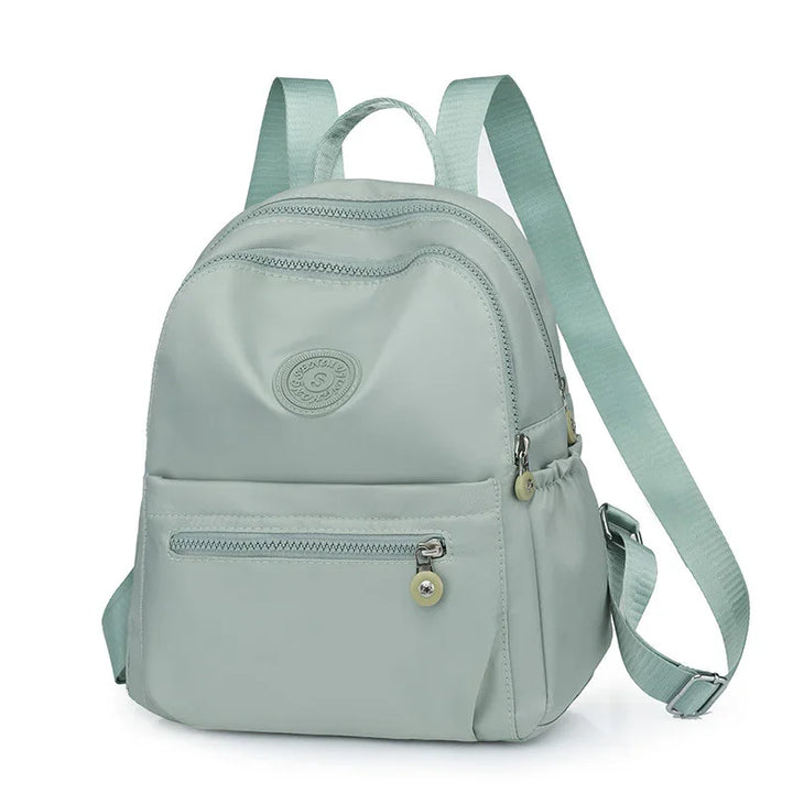 Light gray backpack with front zipper pocket, adjustable straps, and circular logo badge