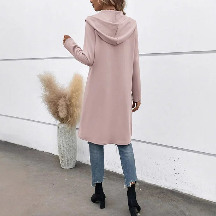 Woman wearing long pink hooded cardigan, cropped blue jeans, and black ankle boots against white wall