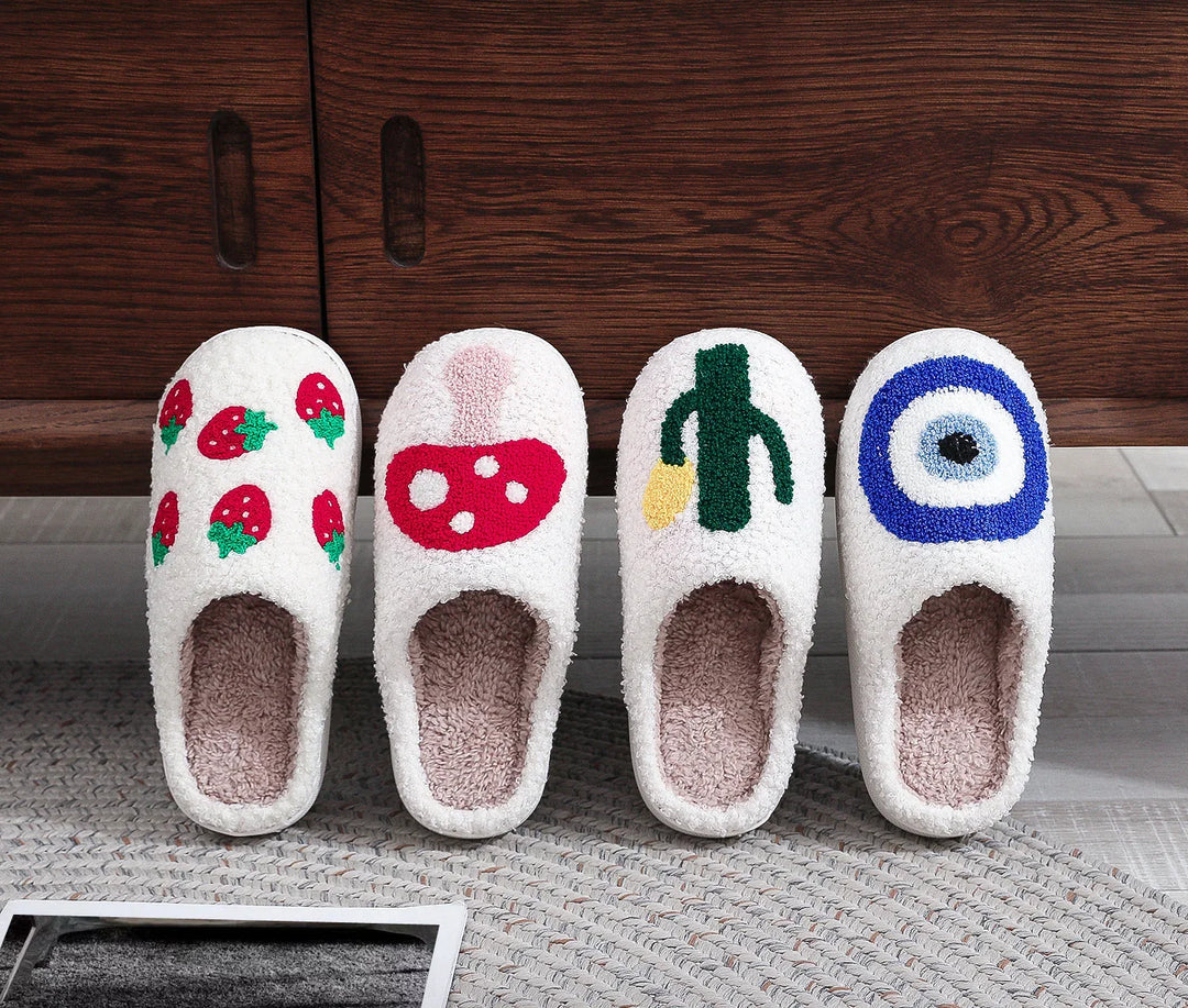 Four pairs of white fluffy slippers with stitched strawberry, mushroom, cactus, and target designs on carpet