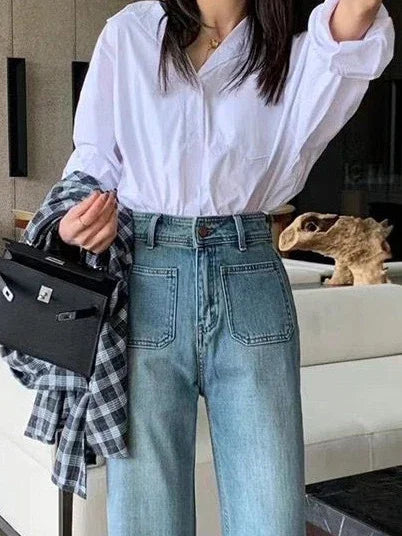 Woman wearing high-waisted blue jeans and a white button-up shirt holding a black handbag and plaid shirt indoors