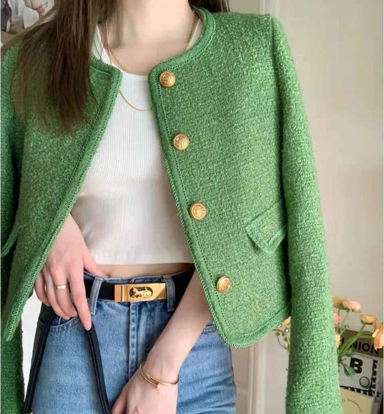 Woman wearing textured green cropped jacket with gold buttons, white crop top, blue jeans, and gold accessories