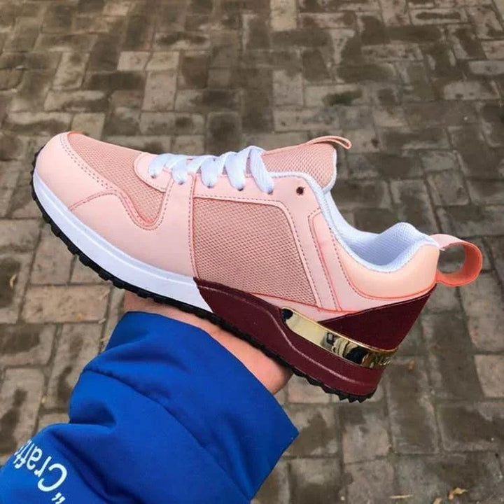 Hand holding pink and maroon athletic sneaker with white laces and gold accent on outdoor brick pavement